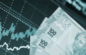 #pracegover Foto mostra três notas de R$ 100 e atrás um quadro do mercado financeiro