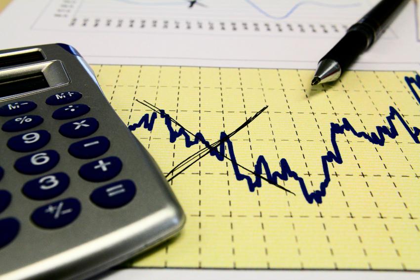 #pracegover Imagem de uma calculadora em cima de um papel com um gráfico oscilando os traços, em alusão à economia do país que vive altos e baixos. Também há uma caneta no canto superior da foto