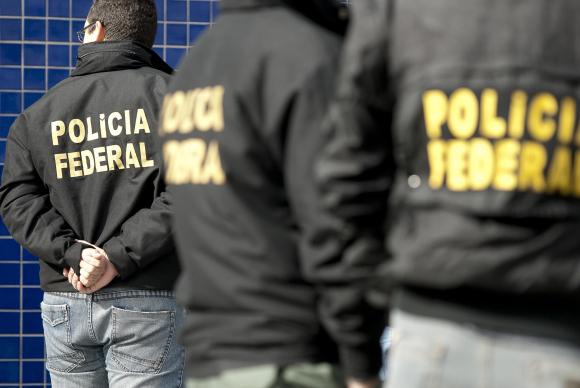#pracegover Policiais federais de costas usando jaquetas de couro pretas com a frase Polícia Federal escrito em letras amarelas.