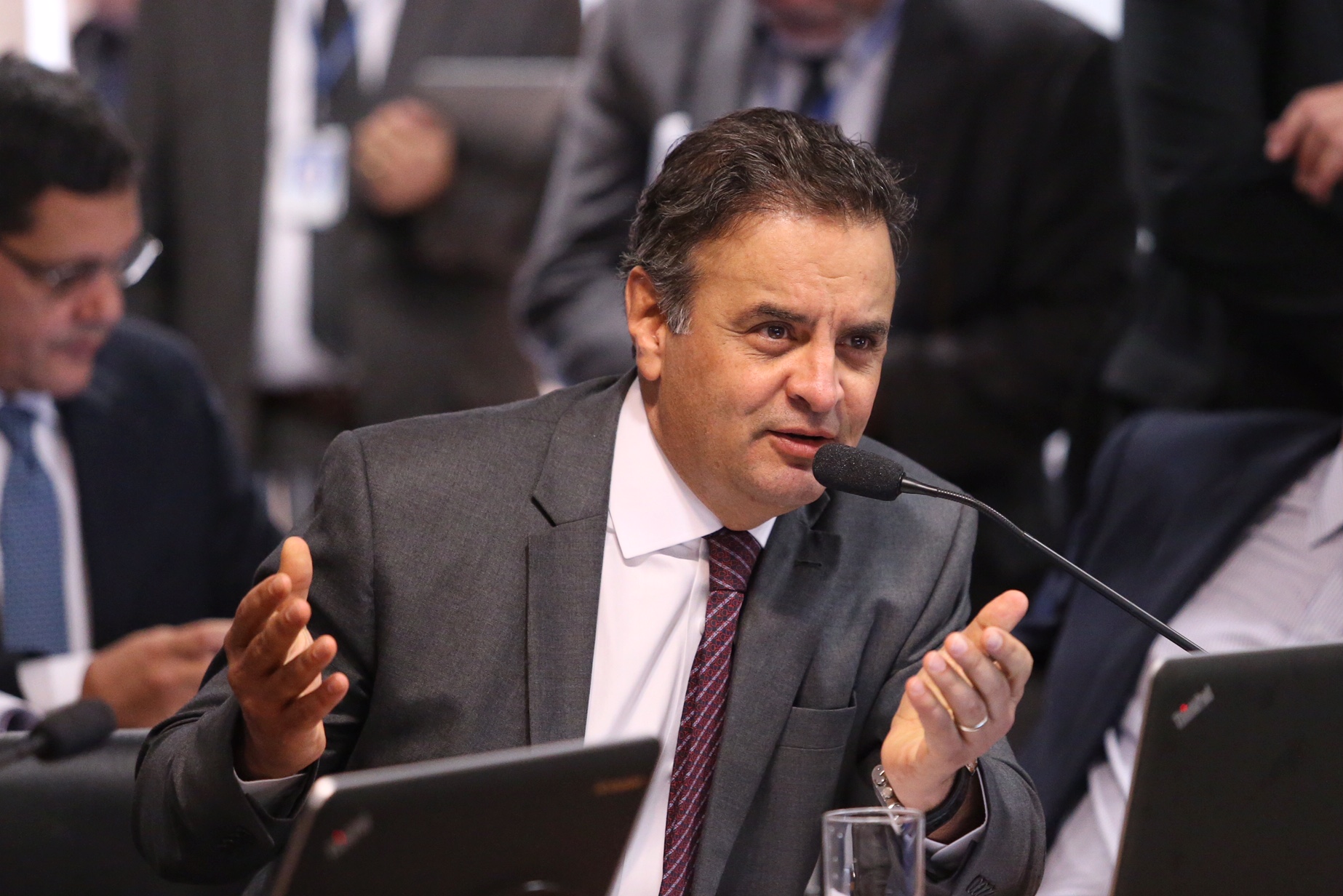 #pracegover senador Aécio Neves na bancada do Senado gesticulando com as mãos na frente do microfone. Ele usa um terno cinza e gravata cor de vinho.