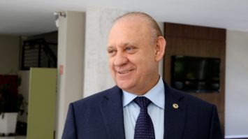#cegopraver: Ademar Traiano está de paletó e gravata azuis e camisa branca em pé, sorrindo e concedendo entrevista. Na foto, aparece apenas uma mão segurando um gravador