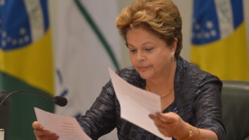 #pracegover: foto mostra Dilma Rousseff com dois papéis nas mãos e bandeira do Brasil atrás