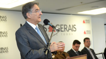 #pracegover: Governador Fernando Pimentel está em pé falando mo microfone no púlpito e atrás o símbolo da bandeira de Minas e outras duas pessoas sentadas