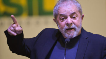#pracegover Ex-presidente Lula está com paletó e blusa azul e com dedo indicador do lado direito apontado