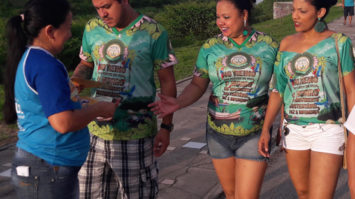 #pracegover: foto aparece três foliões na rua recebendo de um panfleto de uma assistente da prefeitura de Manaus sobre combate ao trabalho infantil e exploração sexual de crianças e adolescentes