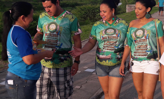#pracegover: foto aparece três foliões na rua recebendo de um panfleto de uma assistente da prefeitura de Manaus sobre combate ao trabalho infantil e exploração sexual de crianças e adolescentes