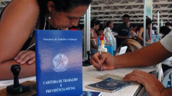 #pracegover: foto mostra carteira de trabalho em destaque em pé em cima de outras empilhadas. Ao fundo, um mulher debruçada em galpão sendo atendida por outra