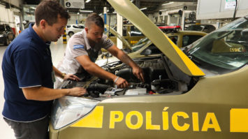 #pracegover: foto mostra carro da polícia militar de Porto Alegre na oficina com capô levantado e mecânicos mexendo