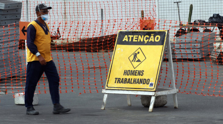 #pracegover: foto mostra obras sendo feitas em aeroporto de Salvador. Tem um operário andando na pista de Salvador e uma placa amarela e preta com "Atenção, homens trabalhando"