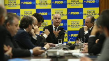 #pracegover: foto mostra bancada de deputados do PSDB sentados em reunião na Câmara e em destaque painel em amarelo e azul com sigla PSDB