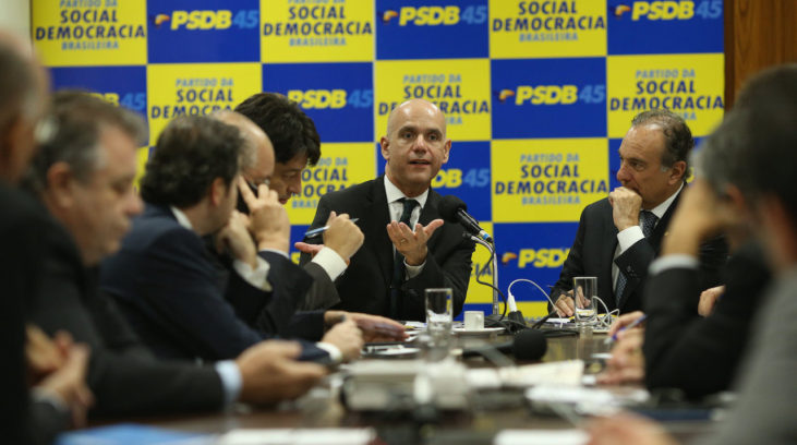 #pracegover: foto mostra bancada de deputados do PSDB sentados em reunião na Câmara e em destaque painel em amarelo e azul com sigla PSDB