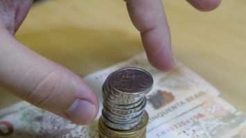 #pracegover: foto mostra uma mão em direção a uma pilha de moedas de R$1 e R$ 0,50 sob duas notas de R$ 50