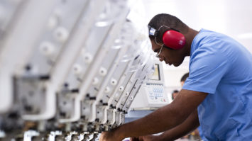 #pracegover: foto mostra operário na linha de produção, usando uniforme azul e EPI de ouvido vermelho