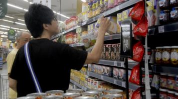 #pracegover: foto mostra mulher vestida de preto de costas escolhendo produto em gôndola do supermercado