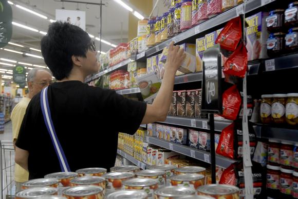 #pracegover: foto mostra mulher vestida de preto de costas escolhendo produto em gôndola do supermercado