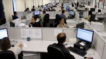 #pracegover: foto panorâmica mostrando pessoas sentadas nos cubículos de trabalho em frente ao computador