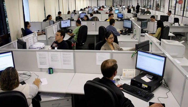 #pracegover: foto panorâmica mostrando pessoas sentadas nos cubículos de trabalho em frente ao computador