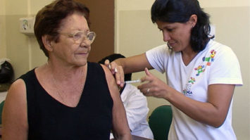 #pracegover: foto mostra senhora vestida de blusa preta e óculos sendo vacinada por enfermeira vestida de branco