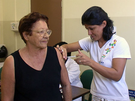#pracegover: foto mostra senhora vestida de blusa preta e óculos sendo vacinada por enfermeira vestida de branco