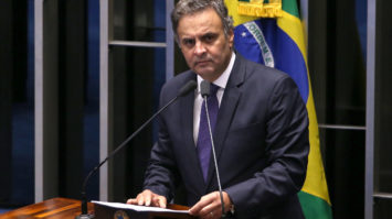 #pracegover: foto mostra senador Aécio no púlpito do Senado falando ao microfone e segurando papeis. Atrás do senador está a bandeira do Brasil no mastro