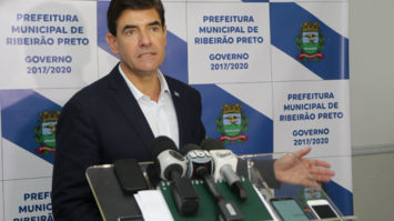 #pracegover: foto mostra prefeito Duarte Nogueira dando coletiva de imprensa. Ele está em pé, na sua frente um púlpito segurando vários microfones e atrás um panei branco e azul
