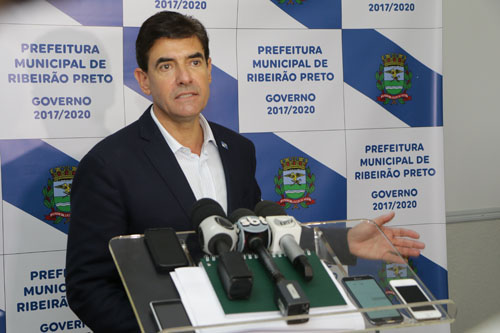 #pracegover: foto mostra prefeito Duarte Nogueira dando coletiva de imprensa. Ele está em pé, na sua frente um púlpito segurando vários microfones e atrás um panei branco e azul
